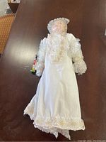 Doll wrapped in bubble wrap on a dark brown table, wearing a white dress with gold floral embroidery, holding a small colorful figurine. Doll arms and head wrapped in bubble wrap.