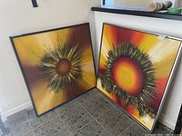 Two abstract sun paintings leaning against a white wall on a tiled floor, showing vibrant yellow, orange, red, and black colors with radiating brush patterns.