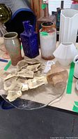 Photo of multiple vases including two Kaiser ceramic vases, a white faceted vase, brown and white glazed jar, and a glass serving plate with oyster shells.