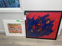Two artworks displayed side by side on the floor: one large framed print with red and blue abstract design, one smaller matted watercolor with colorful abstract shapes including yellow and green forms