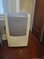 Front view of the Frigidaire portable air conditioner unit placed on a wooden floor near a window and door.