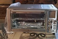 Small stainless steel air fryer oven on a table with front view showing digital control panel, glass door, and internal racks wrapped in plastic.