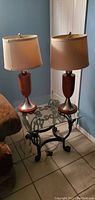 Photo of two similar table lamps showing wooden bodies with sterling steel bases, beige lampshades, placed on a glass and metal table.
