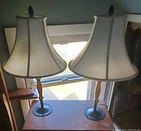 Pair of two cream-colored table lamps with bell-shaped shades on a small wooden table against a window