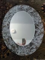 Oval decorative glass mirror with floral patterns on the border, reflecting a candle and a phone, measuring about 23 inches in height.