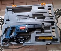 Ryobi 6 amp reciprocating saw in gray molded plastic case with orange latches. Saw is blue and black with electric cord coiled inside. Yellow prying tool and some small accessories placed in molded compartments.