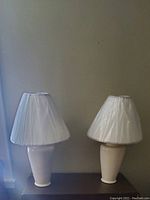Two white ceramic table lamps with fabric shades on a dark surface against a beige wall.
