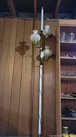 Full view of the vintage gold metal floor lamp with hobnail milk glass shades against wood paneling.