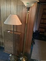 Photo showing two brass-finished floor lamps, one torchiere style and one with pleated fabric shade, standing next to a wooden 3-shelf shoe rack with lock.