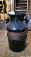 Front view of the vintage metal milk can painted black with twine wrapped handles, showing wear and scratches.