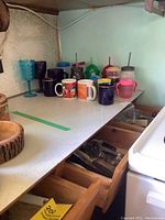 Photo of ceramic mugs, plastic drinking glasses with lids and straws, and kitchen counter with drawers opened showing cooking utensils.