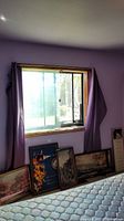 Five framed wall art pieces leaned against a wall beneath a window with purple curtain. Prints include floral art, house or townhouse scenes, wine theme, and horse on bridge. One long narrow plaque with poem and photo is shown upright separately.