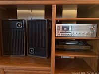 Two wooden framed speakers with black grilles and circular tuning components placed inside a wooden cabinet alongside the stereo receiver and turntable.