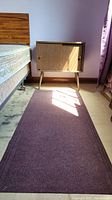 Vintage wood stand beside a bed, showing angled legs, doors, wood grain finish and scratches. Purple floor runner visible on the floor in front of the stand.
