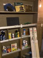 Shelf with multiple household products including sprays, filler powder, pest traps, mini blinds in box, and area rug visible on floor.