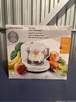 Front view of the product box showing the Baby Brezza One Step Baby Food Maker with fresh vegetables and fruits around, highlighting the one-step cooking and blending feature.