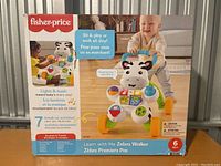 Front view of Fisher-Price Learn with Me Zebra Walker box showing baby playing with the walker and highlighting features such as lights, music, and play activities.