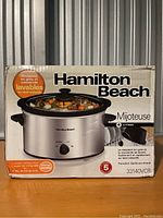 Box of Hamilton Beach slow cooker showing front view with product image and 4-litre capacity, dishwasher safe stoneware, and heating settings features.