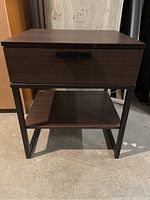 Front view of dark wood IKEA nightstand showing single drawer and open lower shelf, located in basement or storage space.