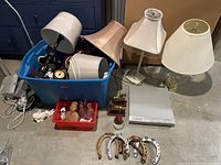Five lamps with various lampshades including white and beige, plus plastic bin containing some items, metal horseshoes, and a silver device.