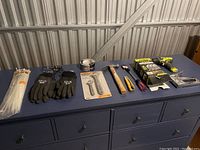 Wide shot showing all assorted tools laid out on a dark blue dresser surface including gloves, hammer, utility knives, tape, wrench set and drill box.
