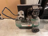 Photo shows side view of green Speedaire compressor unit with handle and large wheels, displaying brand label and wear.