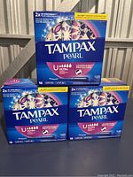 Photo showing three unopened boxes of Tampax Pearl Ultra tampons on a black surface outdoors. The boxes are blue with purple accents.