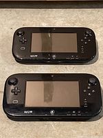 Two black Wii U gamepads side by side on floor surface, front facing showing screens and buttons.