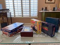 Seven board games stacked and arranged on glass table including Scrabble, Monopoly, The Game of Life, Boggle Jr Letters, Pictionary, Trivial Pursuit, and Bingo set.
