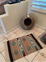 Photo shows decorative clay pot with engraved geometric patterns placed on tiled floor in corner of room beside a small woven rug runner with ethnic pattern in earth tones with fringed edge.
