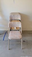 Front view of two stacked padded metal folding chairs showing beige textured cloth covering.