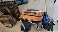Photo showing multiple bags including brown leather-like carry-on bag, green and brown shoulder carry-on bag, textured brown bag, and ski bags in blue and dark colors.