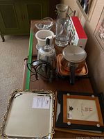 Lot overview showing coffee percolator, fondu pot, Michael Aram tray, Nambe bowl, framed artworks, and various glassware on wooden table.