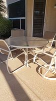 Full view of round outdoor glass top table surrounded by four chairs on patio, two different chair styles shown including swivel and non-swivel.