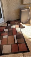 Photo showing two geometric pattern rugs on the ground next to outdoor trash can.