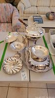 Photo showing full assortment of silver plated serving dishes including covered warming dish with wooden handle, several platters and dishes on table.