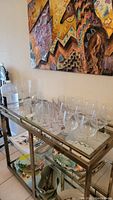 Side angle showing multiple clear glass wine glasses and a pink-fluted glass on a metal glass table with tropical leaf patterned plates on shelf below