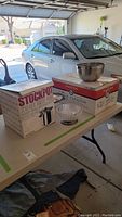 Wide shot on table showing boxed stockpot, boxed chafing dish, stainless steel and glass bowls, and a silver plated serving dish.