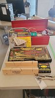 Photo showing red metal toolbox with assorted tools including socket wrench set, screwdrivers, and manual stapler alongside wooden miter box and box of staples.