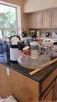 Overview of lot items on a kitchen countertop featuring the air pot, clear bowl with lid, multiple travel mugs and water bottles, and kitchen appliances.