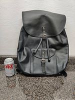 Front view of black leather Helmer backpack with flap and front pocket next to a can of Diet Coke for size reference.