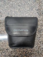Black carrying case with Olympus logo on flap, shown closed on granite countertop.