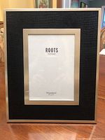 Front view of black fabric photo frame with a silver trim and glass cover, showing the ROOTS Home emblem and photo holder size