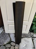 Full view of the black roofing paper roll standing upright against a white door, outdoors on a stone surface.