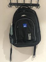 Photo of a black Roots backpack hanging on wall with visible 4 zipper compartments, Roots and AXA logo patches, mesh bottle pocket with bottle, waist and shoulder straps.