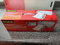 Top view of the unopened Scotch Thermal Laminator packaging showing model TL902-C and product branding in red and white colors, with laminated sheets illustrations.