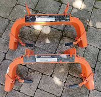 Pair of orange adjustable platform brackets laid on stone surface, showing front with instructional labels and locking mechanisms.