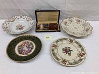Photo showing 4 hand-painted German wall plates with various floral and classical motifs beside a 4-piece steak cutter set in a box
