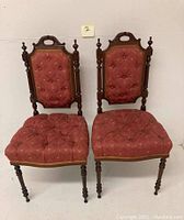 Front view of two Victorian ladies chairs showing tufted reddish fabric upholstery and walnut wood frames with carved detailing and turned finials.