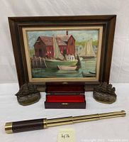 Framed oil on canvas painting showing sailboats and rustic buildings, with metal ship bookends flanking an open wooden box, and a brass telescope placed in front.